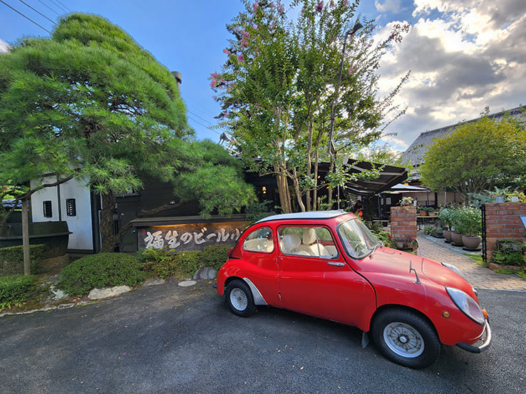 「福生のビール小屋」という木製の看板の前に赤い車（フォルクスワーゲン風）が止まっているアップ写真